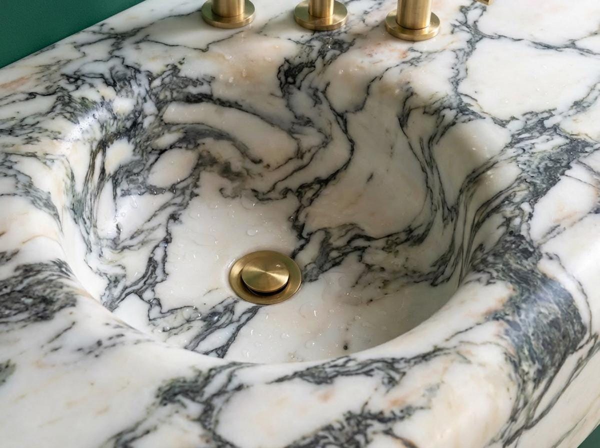 May include: A close-up of a marble sink with a round basin and a gold-colored drain. The sink has a white background with gray and black veining. Three gold-colored faucets are visible in the background.