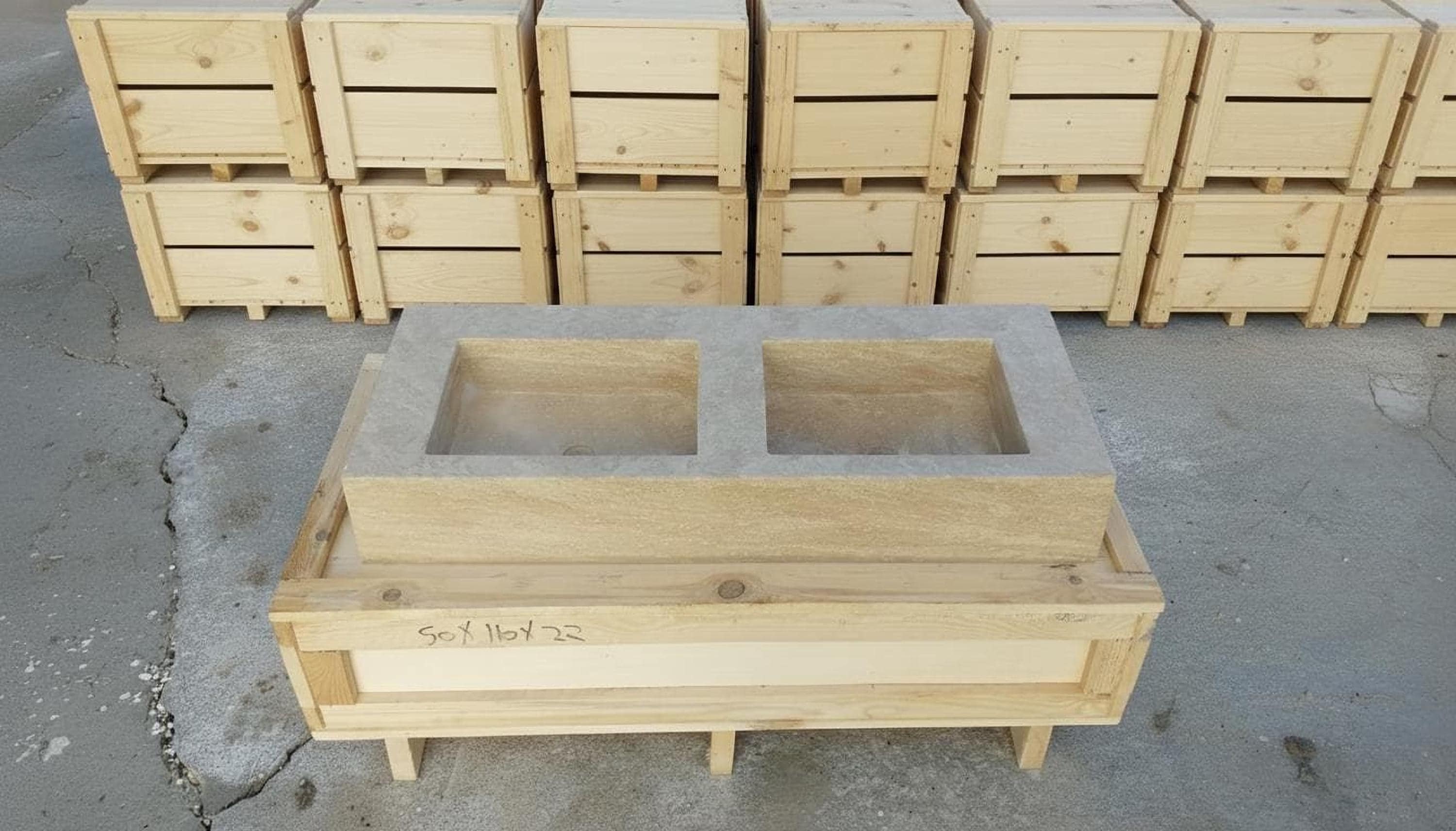 a wooden crate with two rectangular compartments, placed on a concrete floor. Behind the crate, there are several wooden crates stacked on top of each other.