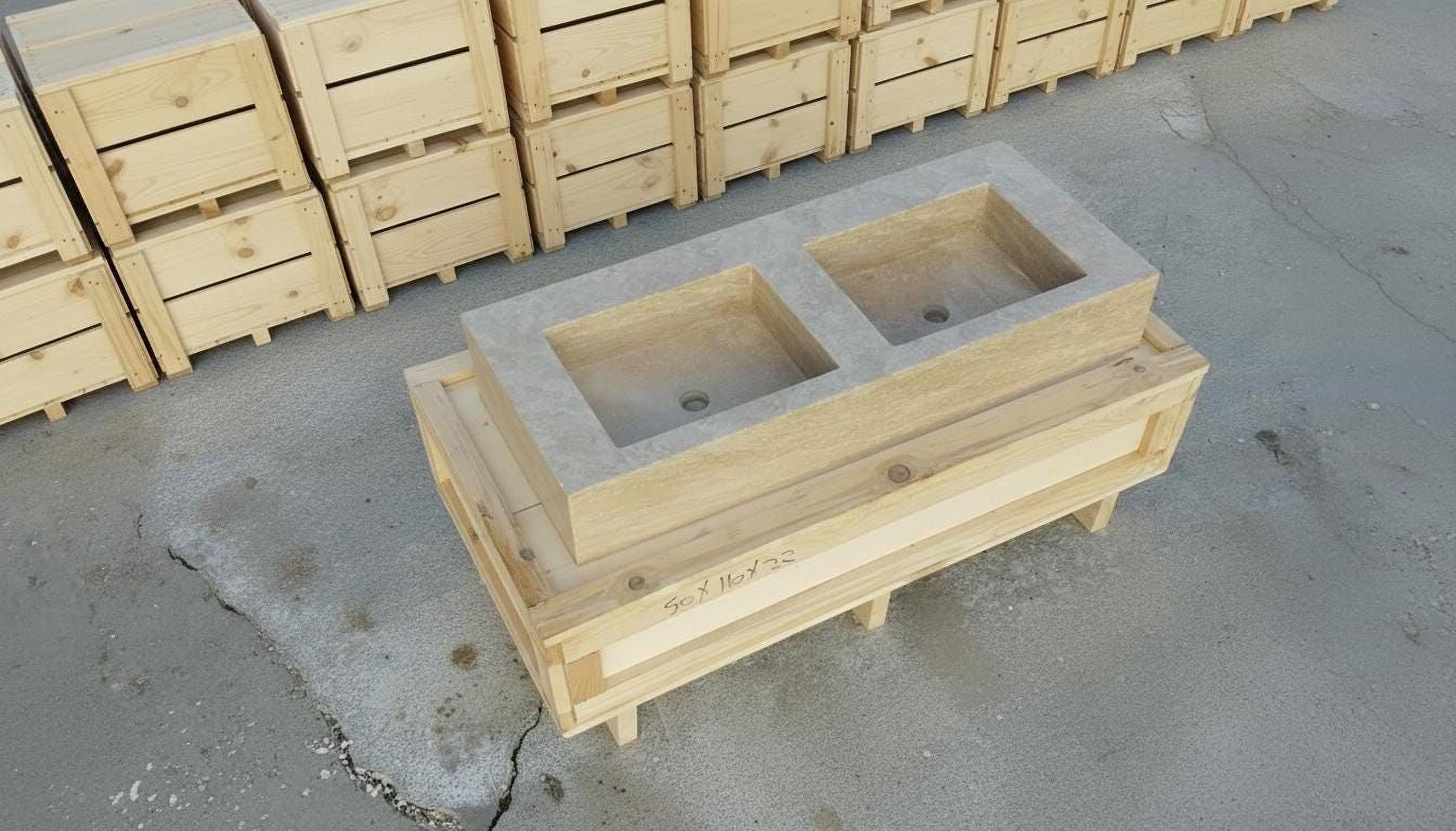 a wooden structure with two rectangular sinks, likely intended for a bathroom or kitchen setting. The structure is placed on a concrete floor, surrounded by several wooden crates or boxes.