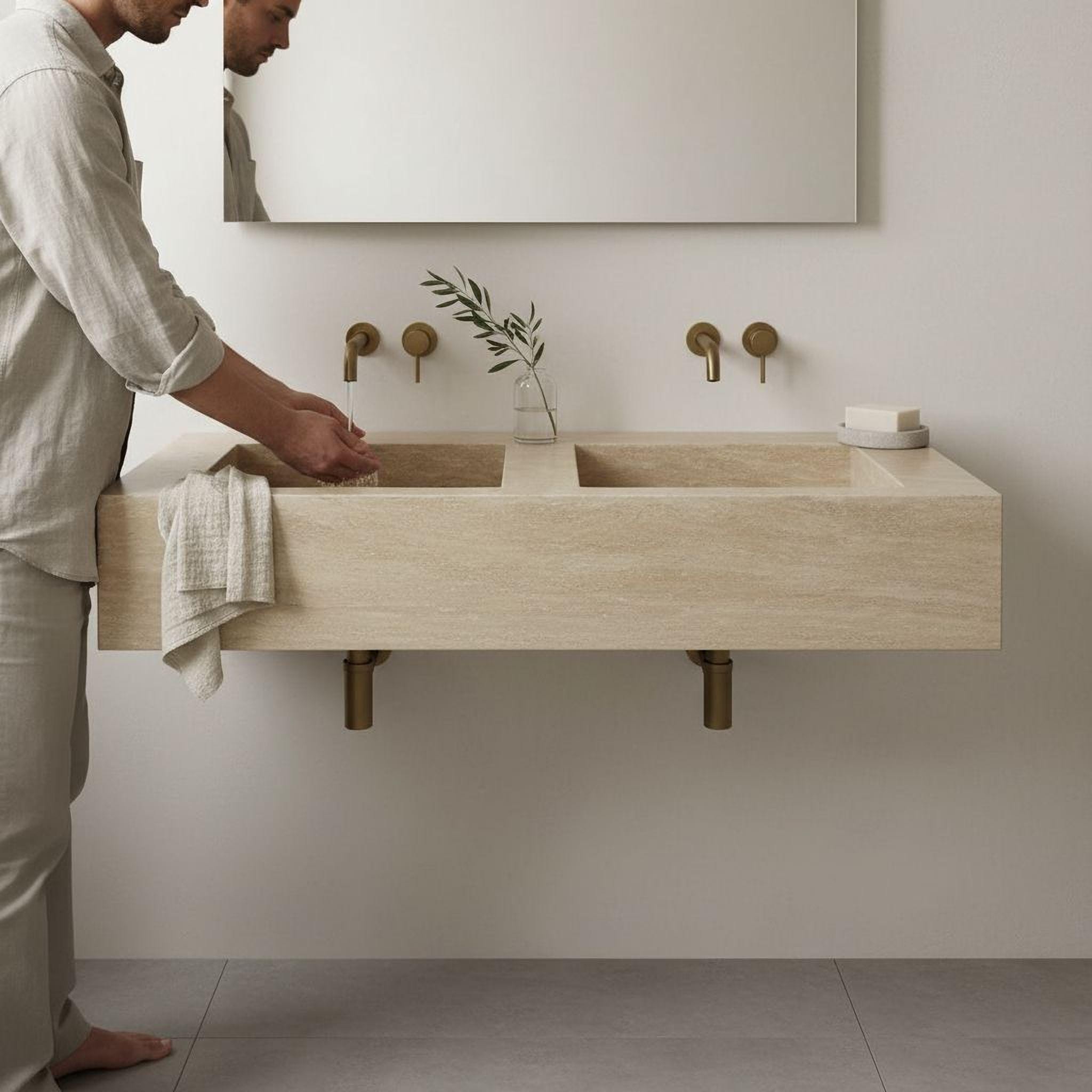A man is standing at a modern bathroom sink, washing his hands. The sink is made of wood and has two faucets, one on each side. The bathroom has a minimalist design with a large mirror above the sink.