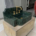 Deep forest-green Verde Marble integrated counter sink with hand-carved vertical reed apron, stepped notched-shoulder backsplash, three-hole brass faucet deck, polished interior, and gold drain fitting, photographed on a wooden crate in a Turkish sto