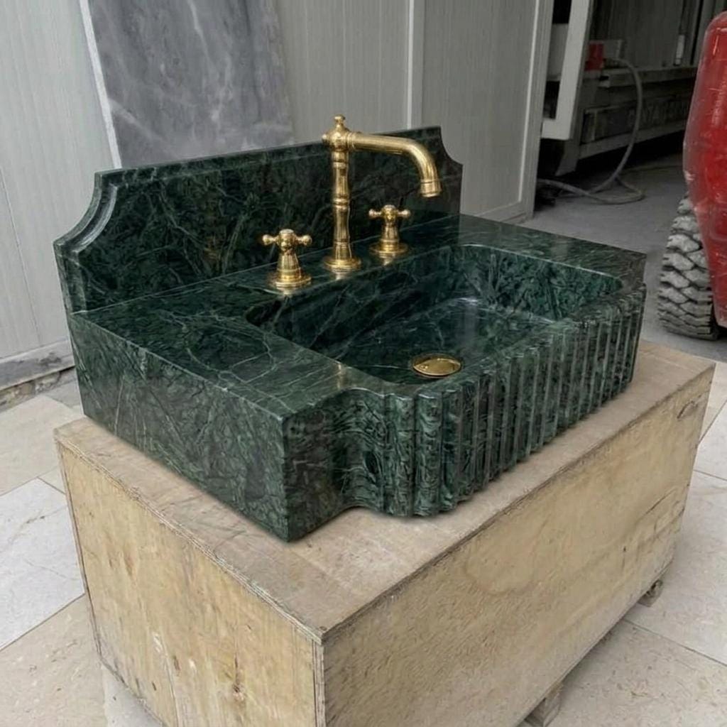 Deep forest-green Verde Marble integrated counter sink with hand-carved vertical reed apron, stepped notched-shoulder backsplash, three-hole brass faucet deck, polished interior, and gold drain fitting, photographed on a wooden crate in a Turkish sto