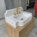 Handcarved Carrara White Marble fluted rectangular sink with integrated counter deck and shaped backsplash, shown with antique brass three-hole faucet, resting on a light oak floating vanity cabinet.