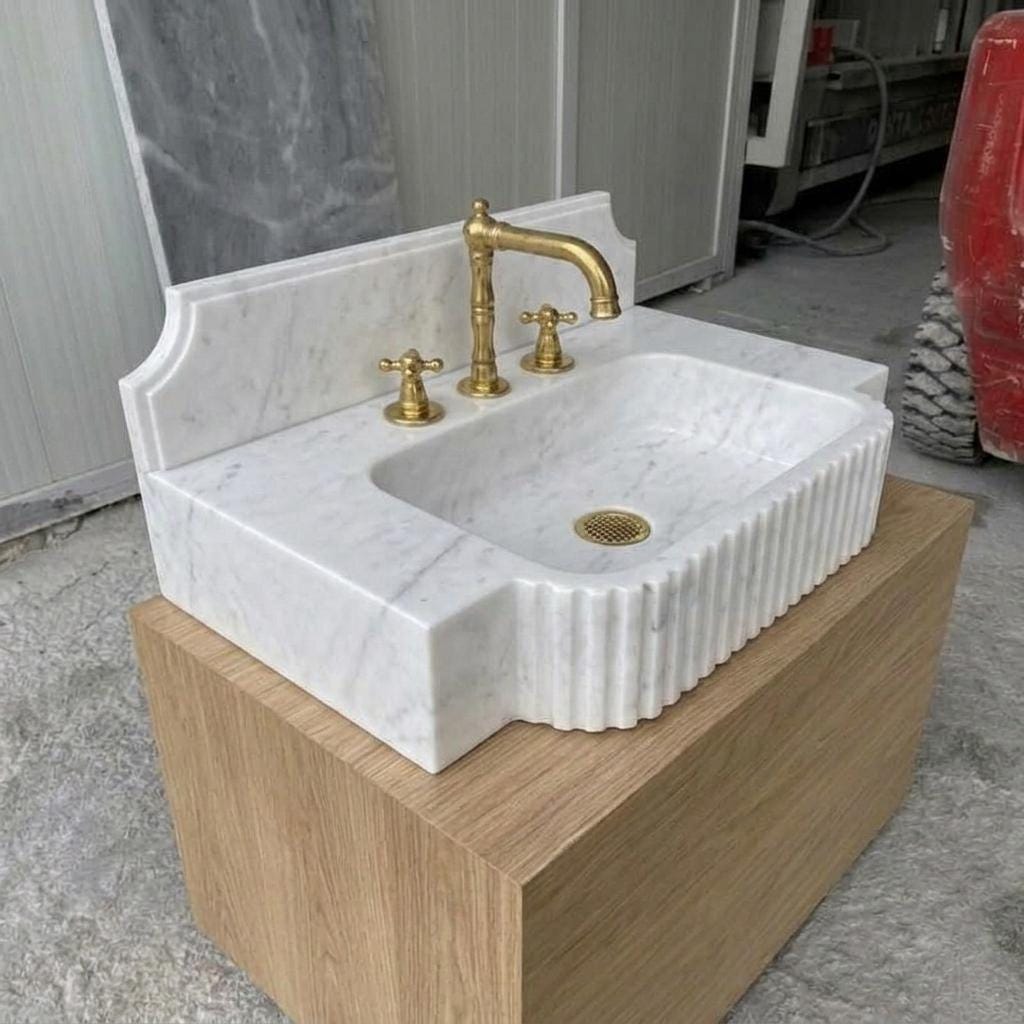 Handcarved Carrara White Marble fluted rectangular sink with integrated counter deck and shaped backsplash, shown with antique brass three-hole faucet, resting on a light oak floating vanity cabinet.