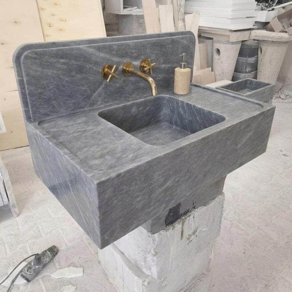 Hand-carved Thundra Gray Marble integrated counter sink with full-width backsplash, showing diagonal white veining across the apron face, a honed matte finish, and a brass cross-handle wall faucet, photographed in a Turkish stone workshop.